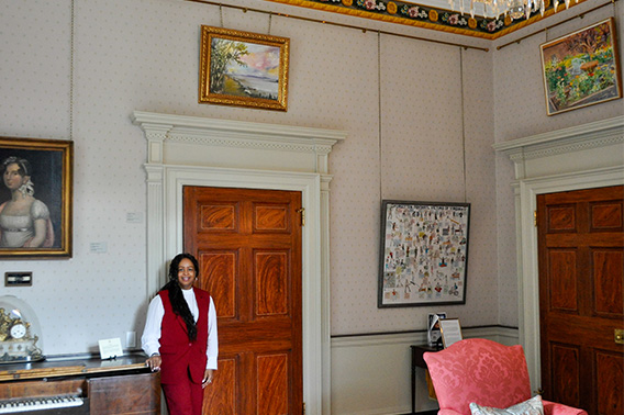 A woman wearing red stands in a well decorated room under a small landscape painting.
