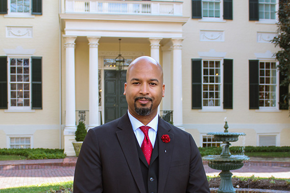 A well dressed man in a three piece suit with a red tie stands in front of a large federal style mansion.