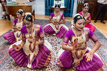 Five women in traditional purple and white attire perform a seated dance on a patterned rug in an elegant room with a piano and blue curtains.
