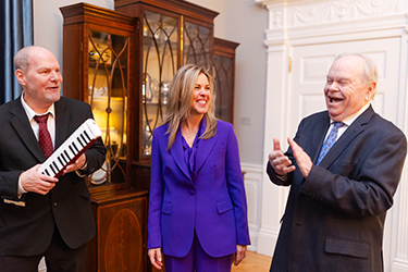 Three people in a formal setting, one holding a small keyboard, another in a purple suit, and an older man laughing.