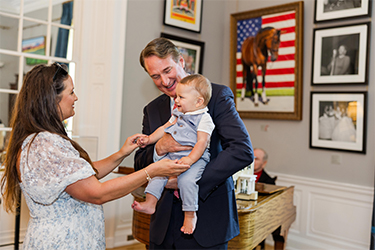 A man holds a baby while standing with a woman in a room decorated with framed artworks.