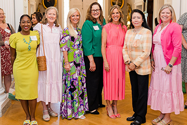 A group of seven women standing in a room, each wearing colorful dresses and smiling.