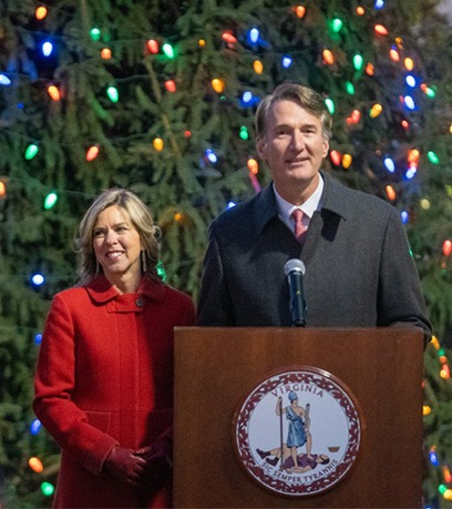 Governor Youngkin and First Lady Suzanne Youngkin stand behing a podium in front of a large Christmas tree with multicolored lights.
