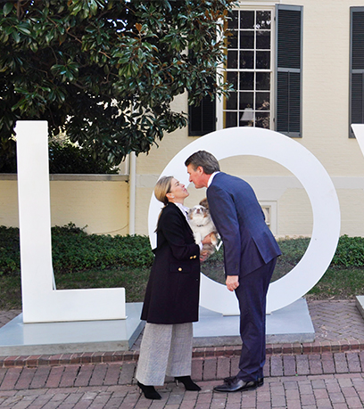 Governor Youngkin and the First Lady lean in for a kiss as they each hold one of their dogs, Bo and Belle, in front of large letters 'L' and 'O'.