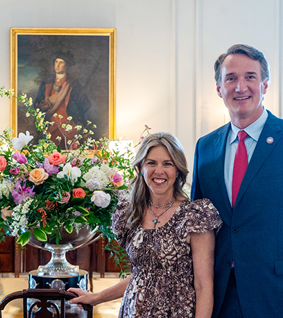 The Governor and First Lady pose in front of the floral arrangement in the Dining Room on Historic Garden Day.