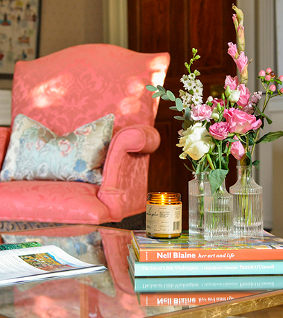 A coffee table with three bud vases with flowers sitting and a candle sitting on top of a stack of books. A coral chair with a decorative pillow is in the background.