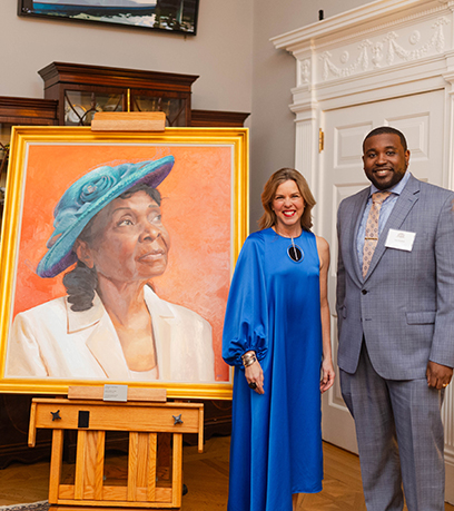 First Lady Suzanne S. Youngkin and artist Stanley Rayfield pose to the right of Stanley's painting, Church Hat No. 32, featuring a woman in a turquoise church hat gazing up and to the right in front of an orange background.