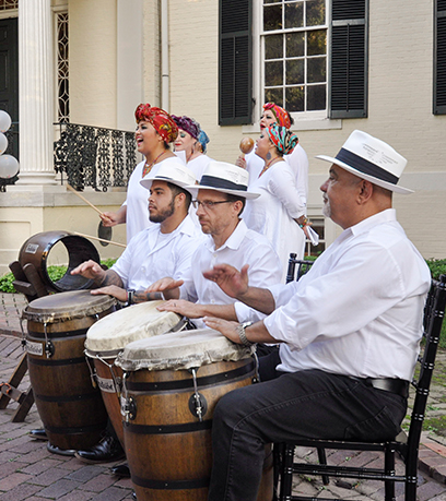 Tradicion Dancy Company performs outside the Executive Mansion. Three men are seated playing drums in the foreground and a group of women sing behind them.