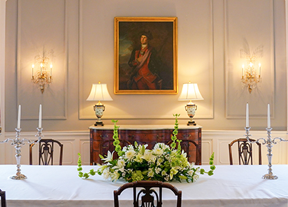 A portrait of a young George Washington hangs in the center of the Dining Room at the Executive Mansion in between two lit lamps and in front of a table with a white cloth and a white and green floral arrangement.