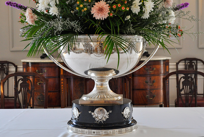 Silver punch bowl filled with floral arrangement resting on white table cloth.