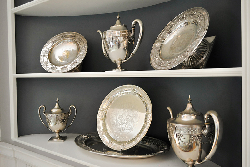 Six pieces of silver (three teapots and three plates) rest on a shelf.